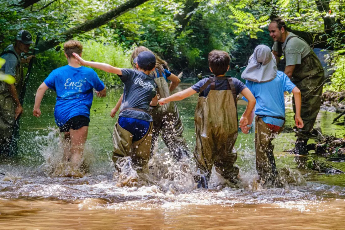 Teens walk in the Eno River, with the water rising above their ankles. Two older instructors stand next to them. They are surrounded by the river's greenery.