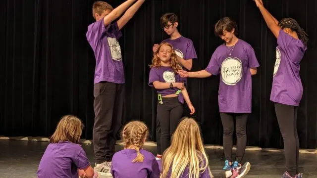 A group of kids wearing purple t-shirts with Vault Theatre logo are participating in a theatre game.