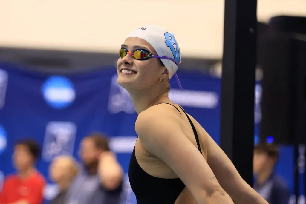 Carolina Swimmer wears a white swim cap with UNC logo, black swimsuit and swim goggles. She is smiling and looking towards the left edge of the picture, audience can be seen in the far background in front of a media backdrop.