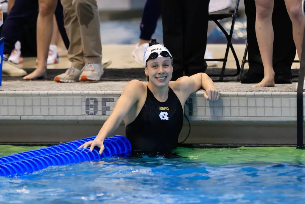 Swimmer in a pool on Carolina's campus with a black swimsuit and swim cap. Swimmer is smiling for the photo with others standing above the pool with just legs visible.