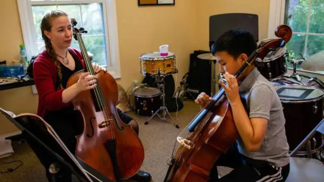 Teacher instructing a student in string intensive camp in a room with windows, both are holding their own bass violin, student is looking down at violin with sheet music placed in front of him while teacher is looking at student.