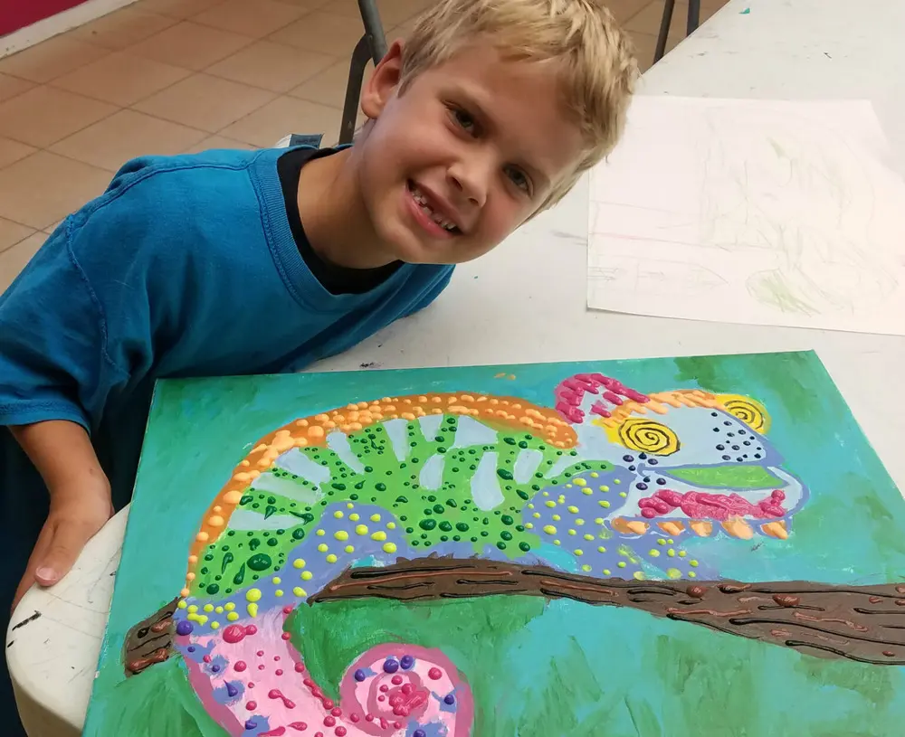 A young child with blonde hair and a blue shirt poses with his colorful artwork of a chameleon on a branch.