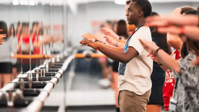 NC State Drum Major student teaching a line of campers in front of a wall of mirrors, students hands are raised as if playing imaginary drums.