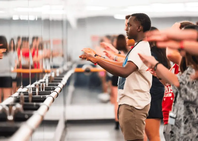 NC State Drum Major student teaching a line of campers in front of a wall of mirrors, students hands are raised as if playing imaginary drums.