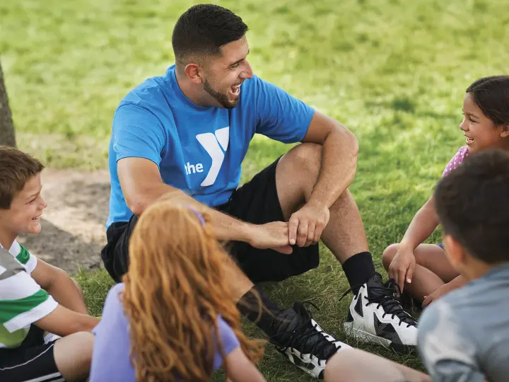 Camp Counselor in a blue shirt sits with YMCA campers in a circle on the grass.