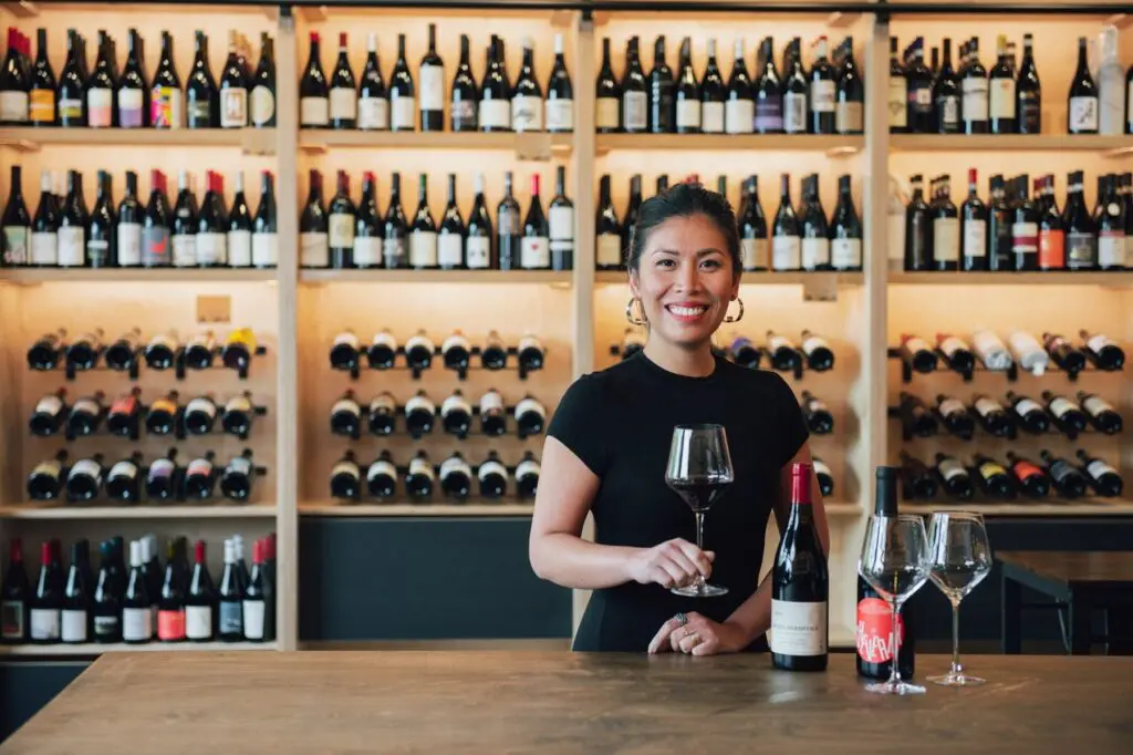 Smiling woman in a black dress standing behind a wood counter with a glass of red wine, in front of backlit shelves filled with wine bottles