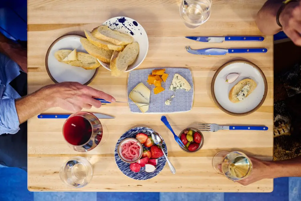 Overhead view of two people sharing a cheese board with sliced baguette, olives, pickled onions, strawberries and glasses of red and white wine on a butcher-block table