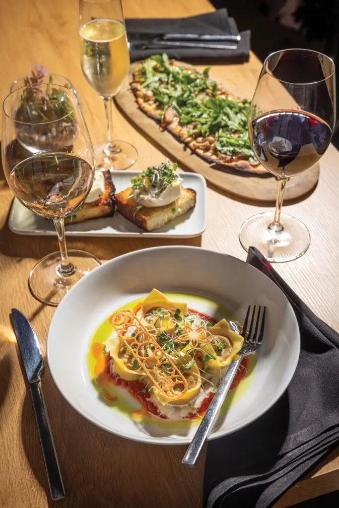 Table set with a bowl of stuffed pasta in tomato and herb oil, a mushroom and arugula flatbread, burrata toast and three glasses of wine