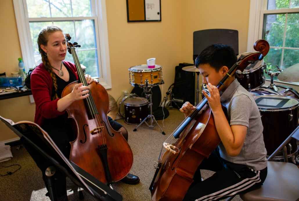 Teacher instructing a student in string intensive camp in a room with windows, both are holding their own bass violin, student is looking down at violin with sheet music placed in front of him while teacher is looking at student.