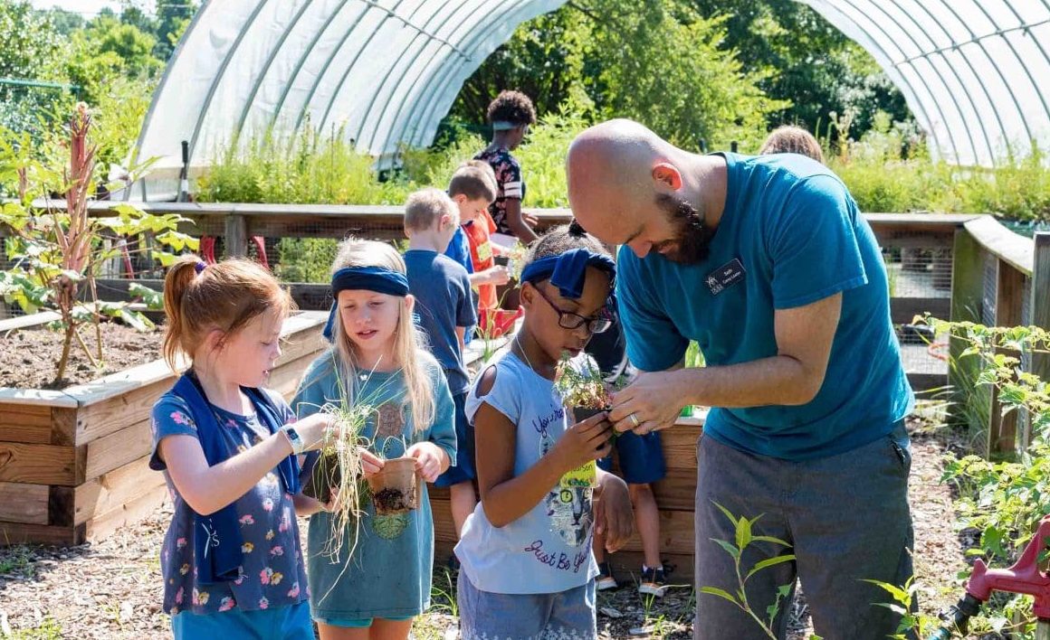 Young campers inside greenhouse at Sarah P. Duke Gardens holding small plants surrounded by greenery. Camp leader in blue shirt looks at a plant held by young female camper.