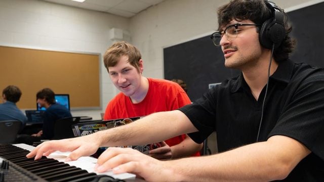 High school students playing the piano with headphones on inside of a classroom with other students.