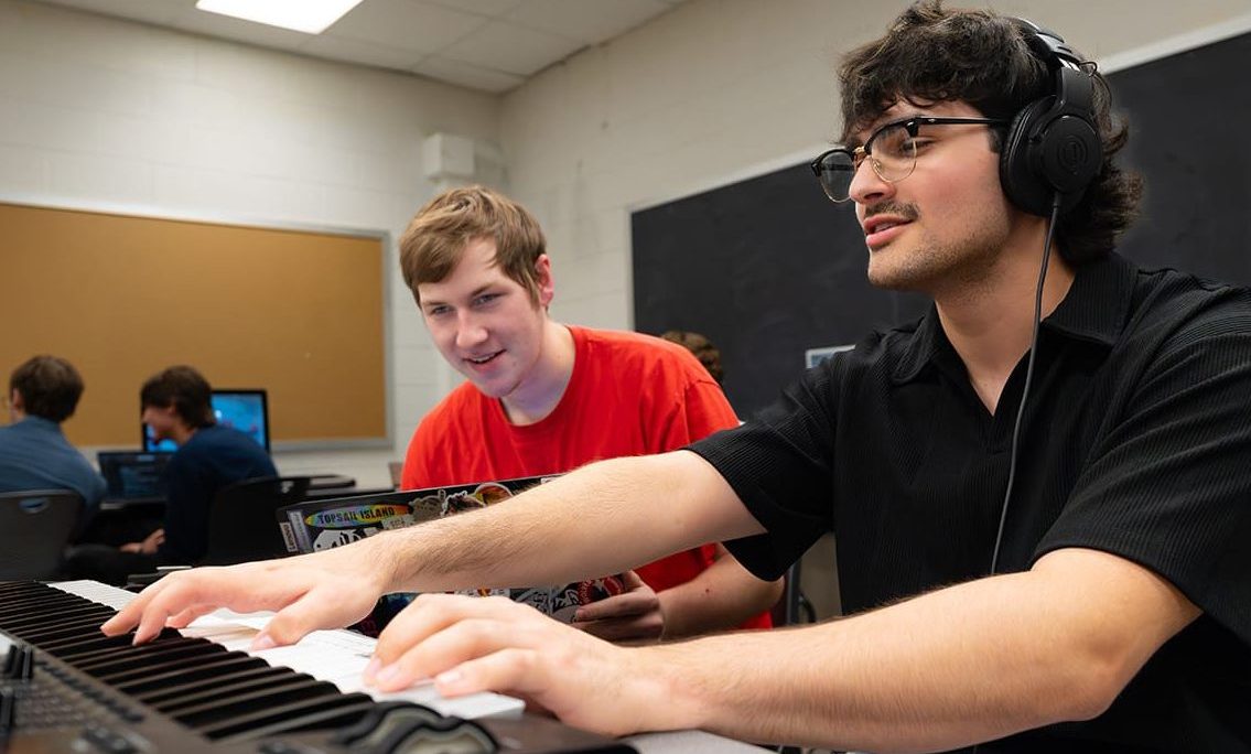 High school students playing the piano with headphones on inside of a classroom with other students.