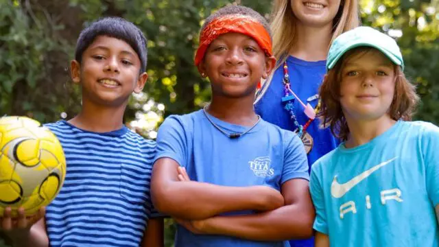Three young campers in blue shirts stand in front of a camp leader at YMCA summer camp. Kid on the far left is holding a yellow soccer ball, trees in the background.